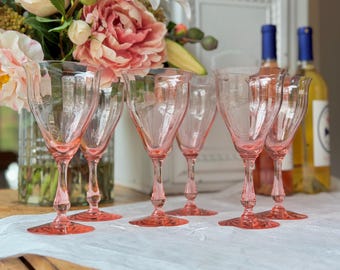 May include: A collection of six pink-tinted wine glasses with a fluted design. The glasses are arranged on a white tablecloth, with a bouquet of pink and white flowers in the background. Two bottles of wine are also visible.