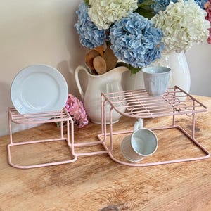 May include: Two light pink metal display stands, one holding a white plate, the other a white cup. A white pitcher with wooden spoons and a vase of blue, white, and pink flowers are in the background. The stands are on a wooden surface.