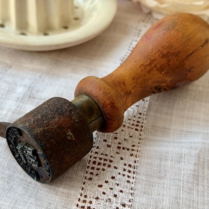 Puede incluir: Sello de madera vintage con un cabezal de sello cilíndrico de metal oxidado. El mango es de color marrón claro y el cabezal del sello tiene un diseño detallado. El sello probablemente se utiliza para manualidades o fines decorativos.