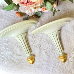 May include: A pair of pale green wall sconces featuring a fluted column design, a wide semi-circular top shelf, and a rounded gold-toned finial at the base. The sconces are constructed with a smooth finish and a classic architectural silhouette.
