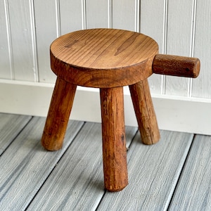 May include: A small, rustic wooden stool with a round seat and three sturdy legs. The stool features a handle on one side. The wood has a warm, natural finish, showcasing the grain. The stool is approximately 10 inches in height.