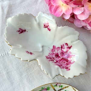 May include: A white, leaf-shaped porcelain dish with pink floral designs and gold trim. The dish features a central depression and is decorated with clusters of pink flowers. A decorative piece for serving or display.