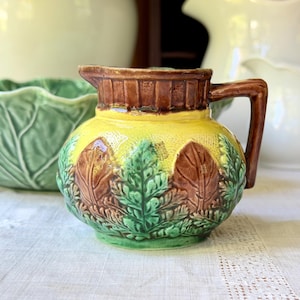 May include: A ceramic pitcher featuring a yellow bulbous body with a textured surface, adorned with raised green and brown leaf motifs around the base. The upper rim is brown with vertical ridges, and it includes a single brown handle.