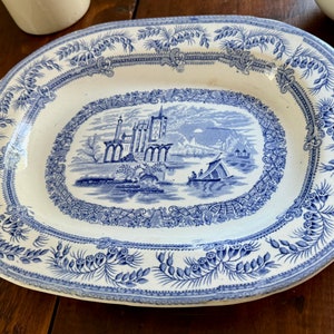 May include: A white oval serving platter with a blue and white floral design around the edge. The center of the platter features a blue and white scene of a castle and a boat on a lake.