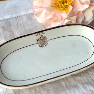 May include: An oval, off-white ceramic dish with a brown rim and a decorative crest. The dish is set on a white linen surface, with a pink and yellow flower in the background. The dish measures approximately 9 inches long.