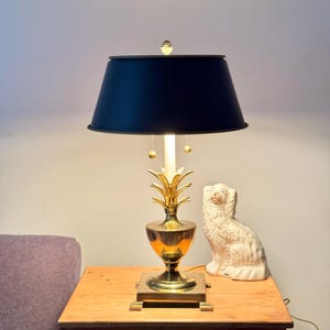 Puede incluir: Una lámpara de mesa de latón con forma de piña y una pantalla azul marino. La lámpara tiene una base cuadrada y una figura de perro de cerámica blanca está al lado. La lámpara está sobre una mesa de madera.