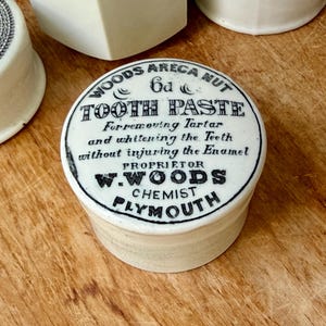 May include: A round white ceramic pot with black printed text that reads: WOODS ARECA NUT 6d TOOTH PASTE For removing Tartar and whitening the Teeth without injuring the Enamel PROPRIETOR W. WOODS CHEMIST PLYMOUTH. The lid features a decorative border.