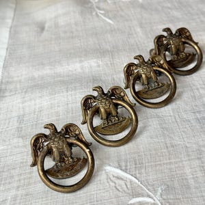 May include: Four antique brass drawer pulls. Each pull features a detailed eagle design with outstretched wings, mounted on a circular ring. The pulls have a vintage, aged appearance, suggesting they are old or have been treated to look old.