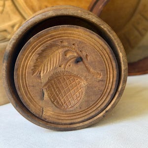 May include: A round, light brown wooden butter mold featuring a detailed carved design of an acorn with a single leaf. The circular stamp is set within a cylindrical wooden frame, showcasing natural wood grain textures and deep, precise carvings.