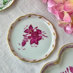 May include: A white, scalloped-edge plate with a floral design. The plate features a magenta flower and leaf pattern with gold trim. The rim has a pink and gold striped border. Other plates are visible in the background.