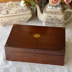 May include: A rectangular wooden box with brass corner accents and a small keyhole. A gold-colored emblem is centered on the lid. The box is set on a white lace tablecloth, with a teapot and flowers in the background.