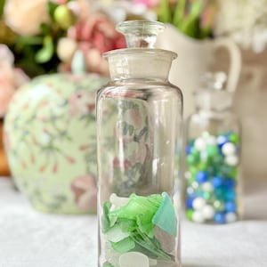 Op de afbeelding: Een heldere glazen apothekersfles met een glazen stop, gevuld met zeeglas in tinten groen, wit en blauw. Op de achtergrond een kleinere pot met blauwe en groene kralen. De potten staan op een wit oppervlak.