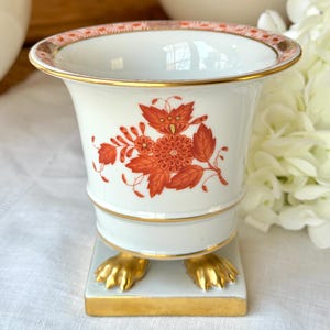 Peut inclure: Un vase en porcelaine blanc avec un dessus évasé et une base carrée. Le vase présente un motif floral dans des tons orange et des accents dorés. La base est soutenue par deux pieds en forme de griffes dorées.