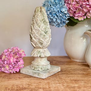 May include: A decorative, weathered-look pineapple sculpture in shades of green and white. The pineapple sits on a square base and has a detailed, textured surface. The sculpture is displayed on a wooden surface, with pink and blue flowers in the background.
