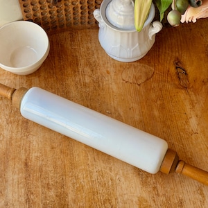 May include: A white rolling pin with wooden handles. The rolling pin is made of a white, opaque material and has wooden handles on each end. The handles are a light brown color. The rolling pin is on a wooden surface.