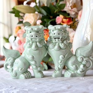 May include: Two celadon-green ceramic guardian lion statues. Each statue has a detailed design with a stylized mane, curled tail, and open mouth. The statues are displayed on a white surface, with flowers and a vase in the background.