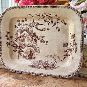 May include: A rectangular, cream-colored serving platter with a brown floral and landscape design. The platter has a decorative border and is part of a matching set. The design includes images of trees, flowers, and a fan.