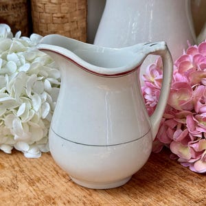 May include: A white ceramic pitcher with a red rim and a black line detail. The pitcher has a curved handle and a rounded base. The background includes white flowers, pink flowers, and a wicker basket.