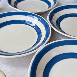 Puede incluir: Un conjunto de platos de cerámica blanca con un diseño de rayas azules. Los platos están dispuestos sobre una superficie blanca, mostrando sus líneas limpias y colores contrastantes. Las rayas azules añaden un toque náutico a la vajilla.