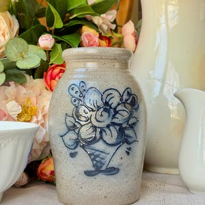 Puede incluir: Un jarrón de gres beige con un diseño floral azul. El jarrón tiene una superficie ligeramente texturizada y una forma cilíndrica. El diseño floral pintado a mano representa un ramo de flores. Es un objeto decorativo.