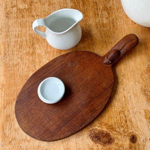 May include: A wooden serving board with a carved handle and a small, white ceramic bowl. A white ceramic creamer pitcher is also present. The board is oval-shaped and has a rich, brown color. The items are arranged on a light-colored wooden surface.