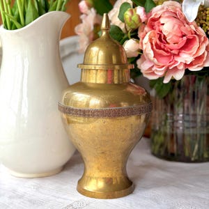 May include: A gold-colored, lidded urn with a decorative band around the middle. The urn is displayed on a white tablecloth, with a white pitcher and a vase of pink flowers in the background. The urn has a classic shape.