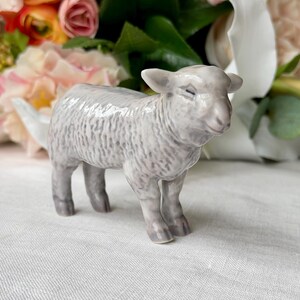 May include: A ceramic lamb figurine in shades of gray and white. The lamb has a detailed wool texture and is standing on a white surface. The background features a bouquet of flowers.