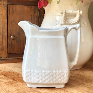 Könnte beinhalten: Eine weiße Keramikkrug mit einem Korbgeflechtmuster am unteren Teil. Der Krug hat einen Griff und einen Ausguss. Eine große cremefarbene Vase befindet sich im Hintergrund. Die Objekte stehen auf einer Holzoberfläche.