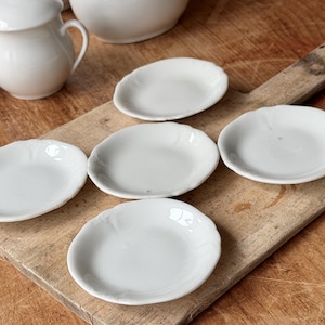 May include: A collection of white ceramic dishes, including small plates with scalloped edges and a creamer pitcher. The plates are arranged on a wooden cutting board, showcasing a rustic aesthetic. The dishes are clean and simple.