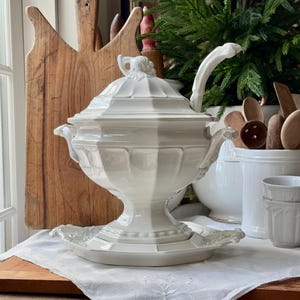 Puede incluir: Una gran sopera de porcelana blanca con tapa y cucharón. La sopera tiene una base decorativa y un asa a cada lado. Está sentada sobre una fuente blanca con un borde decorativo.