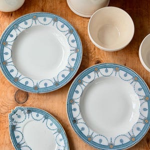 May include: Overhead shot of white ceramic plates and bowls with a blue and gold decorative border. The plates are arranged on a wooden surface, showcasing a classic tableware design. The set includes various sizes and shapes.