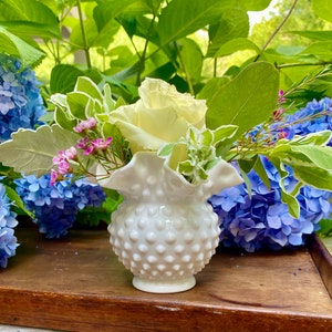 May include: A white hobnail vase holds a cream-colored rose, small pink flowers, and green foliage. The vase is set against a backdrop of blue hydrangeas and green leaves, creating a natural, floral arrangement.
