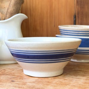 May include: A cream-colored ceramic bowl featuring three horizontal blue stripes around the exterior. The bowl has a wide, flared rim and a tapered base, with a speckled texture visible across the off-white surface of the stoneware material.