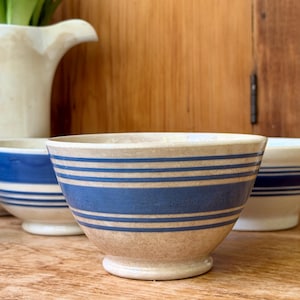 May include: A beige ceramic bowl featuring a series of horizontal blue stripes of varying widths around the exterior. The bowl has a rounded body and a small circular base. It is positioned in front of other similar bowls and a white pitcher.