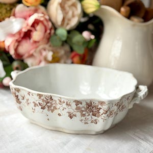 May include: An antique white ceramic serving bowl with a brown floral pattern. The oval-shaped bowl has two handles and a scalloped edge. A white pitcher and flowers are in the background.