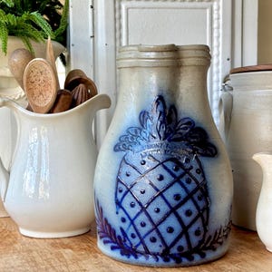 May include: A vintage stoneware pitcher with a blue pineapple design. The pitcher has a light blue background with a dark blue pineapple motif and the text "UNION PORCELAIN WORKS, NEW YORK". Other white ceramic pitchers and wooden utensils are also visible.