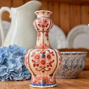 May include: A decorative ceramic vase with a cream-colored base and intricate floral patterns in red, blue, and gold. The vase has a bulbous body and a slender neck, standing approximately 8 inches tall. The design is reminiscent of vintage or antique pottery.