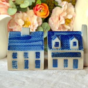 May include: Two ceramic house figurines with blue roofs and white walls. The houses have detailed blue window and door designs. The background includes a white pitcher and a bouquet of pink and orange flowers.