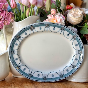 May include: An oval, white serving platter with a teal and dark blue decorative border. The border features a dotted line, floral medallions, and a beaded chain design. The platter is surrounded by white pitchers and fresh flowers.