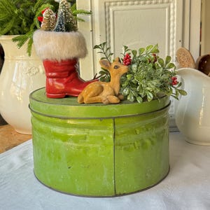 Pode incluir: Uma exibição festiva de Natal com uma bota vermelha brilhante com acabamento em pele sintética, segurando árvores de Natal em miniatura. Uma pequena figura de veado castanho claro repousa ao lado de vegetação e bagas vermelhas em um recipiente redondo de metal verde.