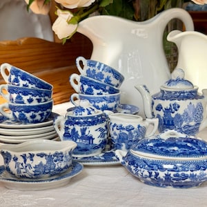 May include: A blue and white porcelain tea set featuring a willow pattern design. The set includes a teapot with a lid, a lidded sugar bowl, a creamer, a gravy boat on a saucer, a stack of saucers, and several teacups with matching saucers.