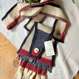 May include: A handcrafted wool scarf with a unique design. The scarf features cream, navy, red, and brown stripes, with a fringed edge. It has a button closure and beaded accents. The scarf is displayed on a white embroidered cloth.