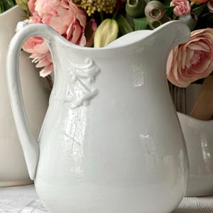 May include: A white ceramic pitcher with a curved handle and a decorative leaf design. The pitcher is set against a backdrop of pink and white flowers, suggesting a farmhouse or vintage aesthetic.