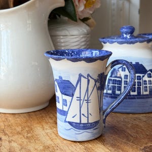 Może przedstawiać: Niebiesko-biały ceramiczny kubek z motywem marynistycznym. Przedstawia żaglówkę i domy. Brzeg jest niebieski, falisty. Widoczny jest pasujący słoik z pokrywką. Umieszczony na drewnianej powierzchni.