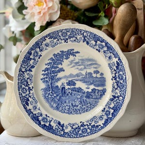 Puede incluir: Un plato decorativo azul y blanco con una escena de paisaje en el centro. El plato tiene un borde festoneado y un borde floral. Está rodeado por una jarra blanca y flores.