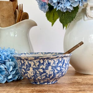 Pode incluir: Uma tigela de cerâmica com padrão azul e branco com uma colher de pau dentro. A tigela está sobre uma superfície de madeira, com um jarro e um vaso brancos ao fundo. Flores azuis e rosas também são visíveis.