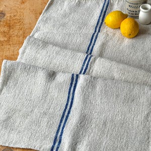 May include: A linen table runner with two vertical blue stripes. Two yellow lemons and a small ceramic jar are on the wooden surface. The runner is made of a light-colored, textured fabric.