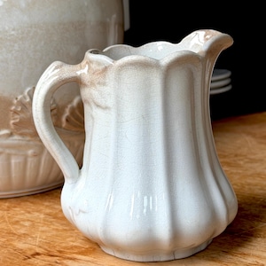 May include: A white ceramic cream jug with a scalloped rim and a vertical ribbed body. The piece features a glossy glaze with a visible network of fine cracks throughout the surface and light brown staining along the handle and top edge.