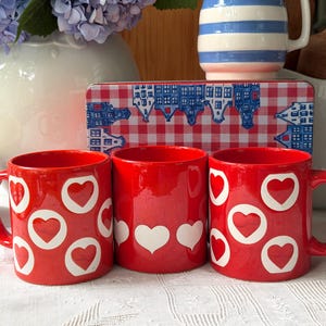 May include: Three bright red ceramic mugs featuring white heart patterns. Two mugs display an all-over heart print, while the centre mug features a single horizontal row of white hearts around the base. The mugs have a glossy finish and curved handles.
