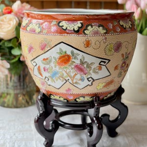 May include: A decorative ceramic planter with a floral design, resting on a dark wooden stand. The planter features a peach-colored background with colorful flowers and a red and gold rim. The stand has a round base and three curved legs.